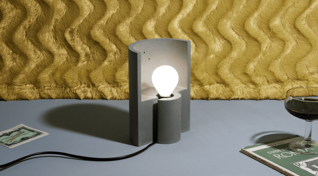 Half-cylinder concrete table lamp, switched on in front of a wavy yellow wall