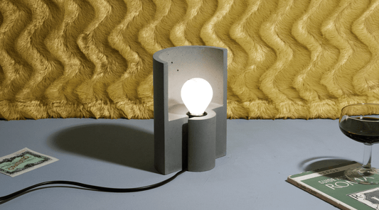 Half-cylinder concrete table lamp, switched on in front of a wavy yellow wall