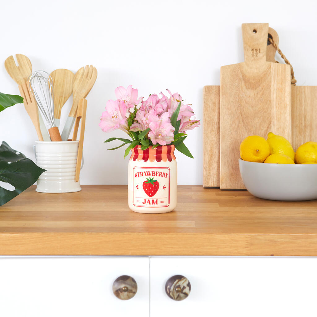 Jam vase, strawberry version with pink flowers, on a worktop between chopping boards and lemons