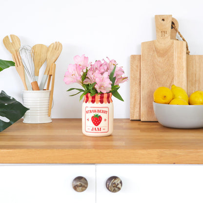 Jam vase, strawberry version with pink flowers, on a worktop between chopping boards and lemons