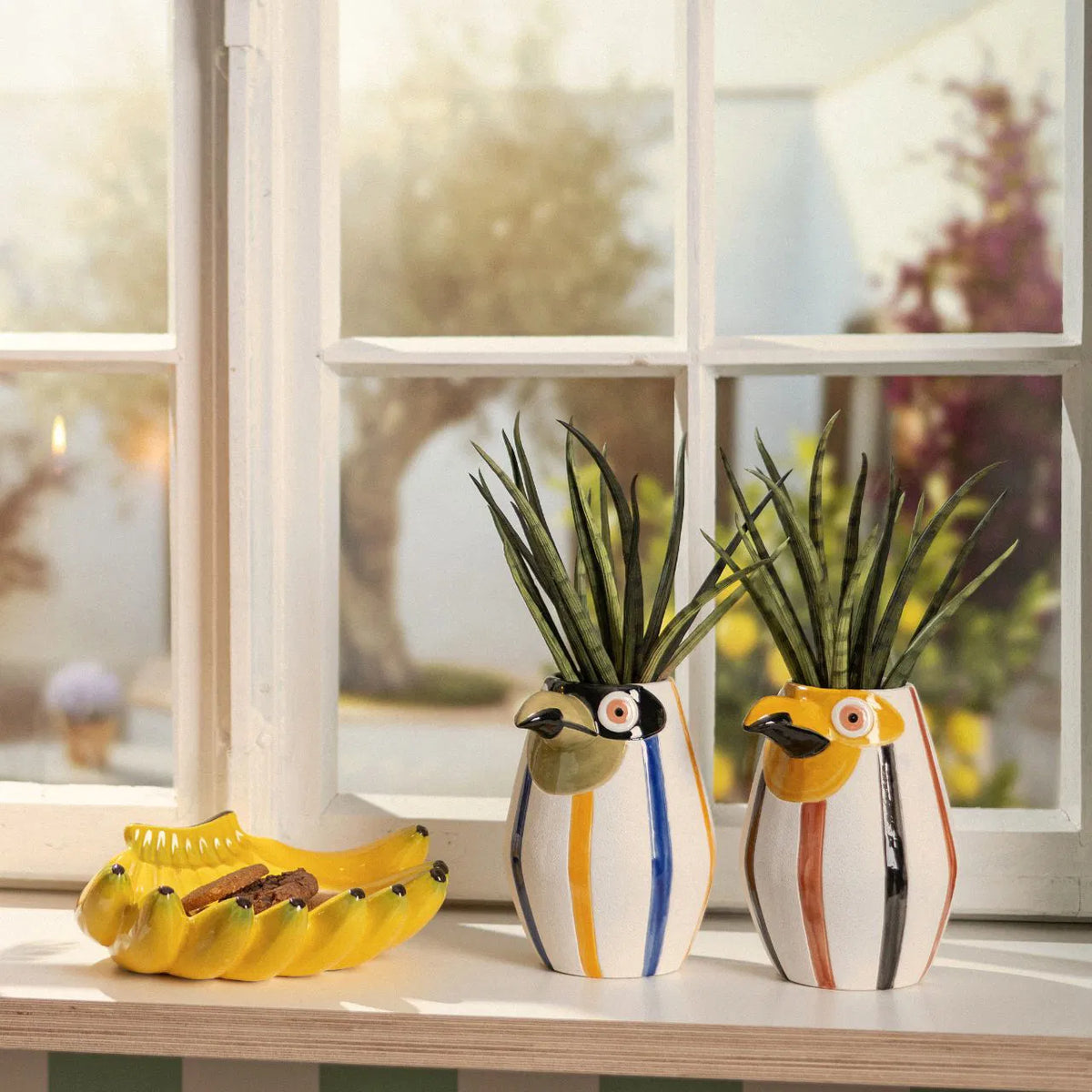 Banana bowl in yellow on a windowsill, among bird-shaped vases with plants.