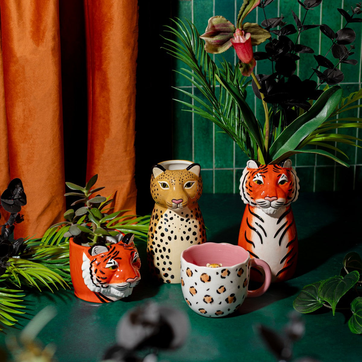 Leopardo vase alongside a tiger-shaped vase and a striped mug, among plants on a green background.
