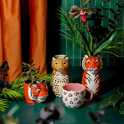 Leopardo vase alongside a tiger-shaped vase and a striped mug, among plants on a green background.