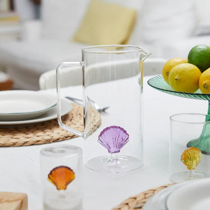 Caraffa Atlantis glass jug with a purple shell inside, on a table set with fruit and glasses.