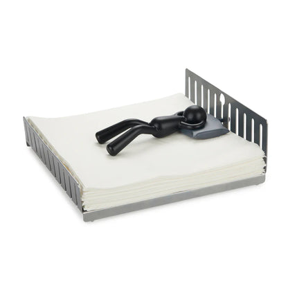 Napkin holder in grey metal, with a small sleeping figure lying on top of the napkins.