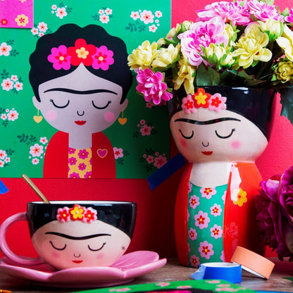 Vaso Frida accanto a una tazza coordinata e a un poster con quel volto, tra fiori colorati.