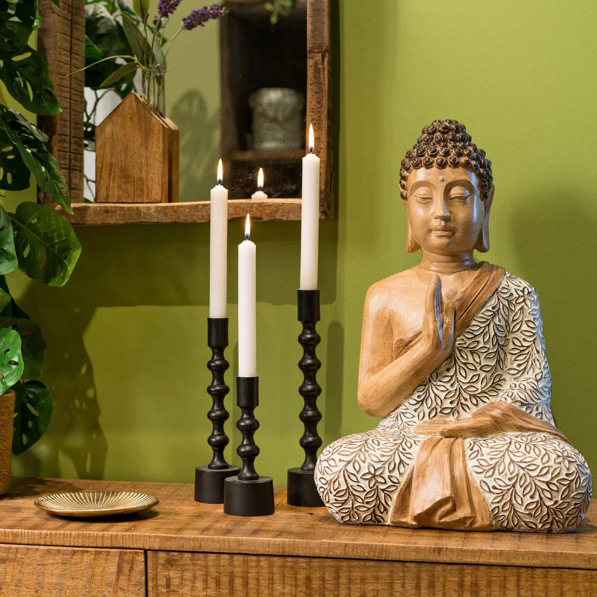 Three Totem candlesticks of different heights with lit white candles, next to a Buddha statue against a green wall.