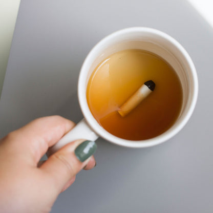 A hand holds a mug containing a cigarette shape and amber liquid