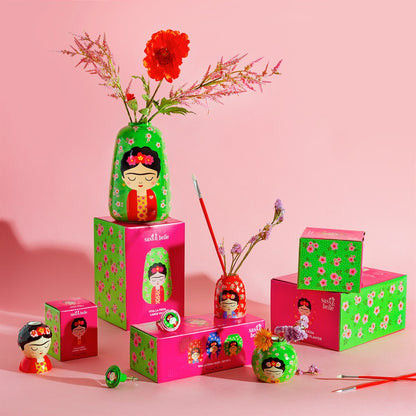 Vaso Frida Floral vases and their boxes scattered on a pink background, with a red poppy and brushes.