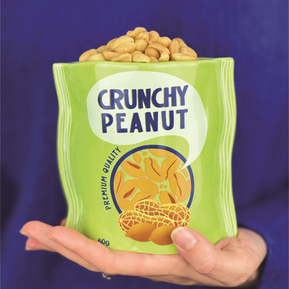 Crunchy Peanut vase shaped like a green bag, held in one hand against a purple background.