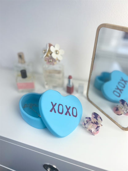 XOXO heart box in light blue, open on a dressing table among perfumes and a mirror.