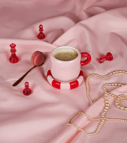 The pink mug on pink bedsheets, with chess pieces, a macaron and a pearl necklace