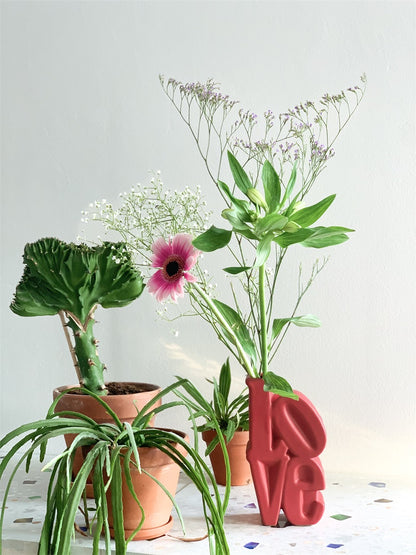 Love vase in red among potted plants and cut flowers, on a mosaic floor.