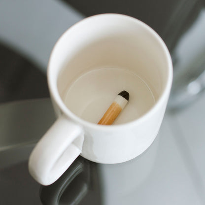 Cigarette Butt mug resting on a glass table, a hand nearby and the fake butt inside.