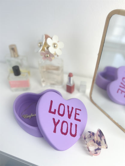 Love You heart box in lilac, open on a dressing table among perfumes and a mirror.