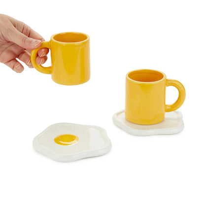 Sunny Side Up coffee cup a hand lifting one yellow mug, with egg-shaped saucers behind it.