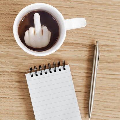 Middle Finger mug white mug seen from above, a middle finger inside the coffee next to a notepad.