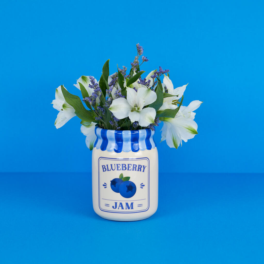Jam vase, blueberry version with white and green flowers, among scattered blueberries on blue