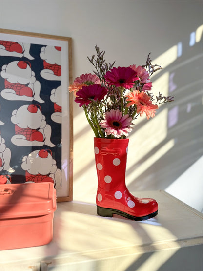 Wellington Pois boot vase in red with mixed pink flowers, on a shelf next to a poster.