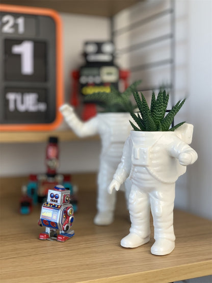 Astronauta vase on a wooden shelf, with toy robots and a calendar in the background.