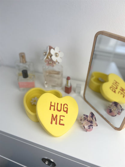 Hug Me heart box in yellow, open on a dressing table among perfumes and a mirror.