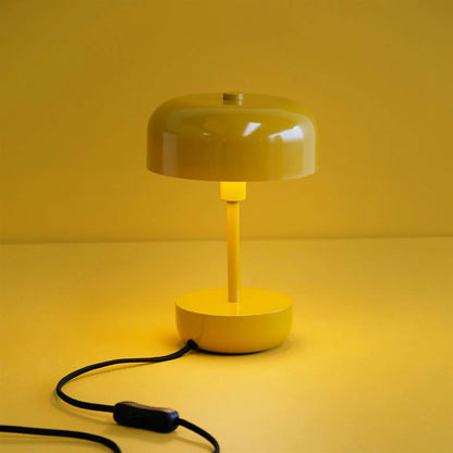 Haipot lamp in mustard yellow, lit, on a yellow background with the black cord on the floor.