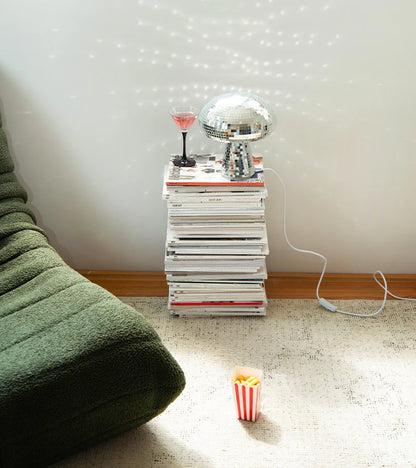 Disco mushroom lamp, mirrored and lit, standing on a stack of magazines beside a green sofa