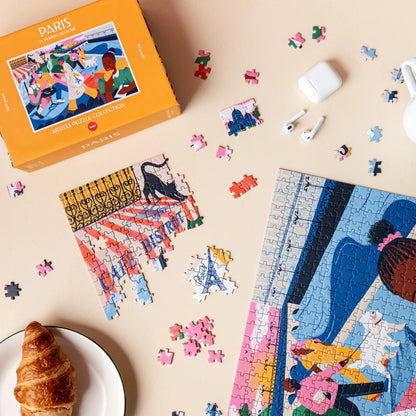 Paris Puzzle box on a table surrounded by scattered pieces, earbuds and a croissant.