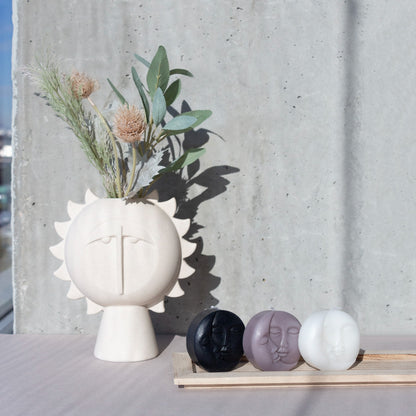 Sun-face vase with dried branches, next to three small faces in black, mauve and white