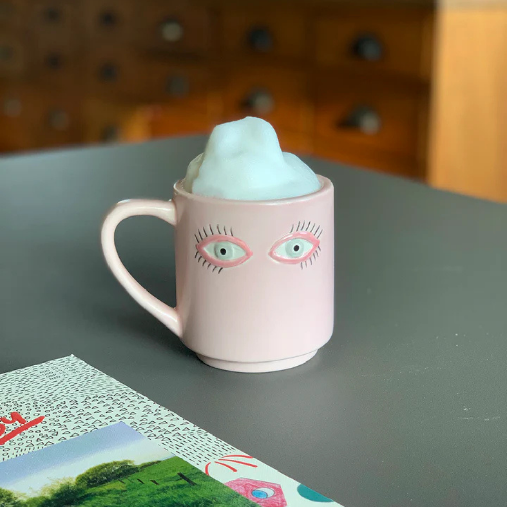 Surreali mugs, pink mug with painted-on eyes, filled with a fluffy blue foam.
