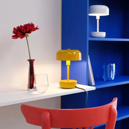 Haipot lamp in yellow, lit on a white desk, with a red flower in a small vase.