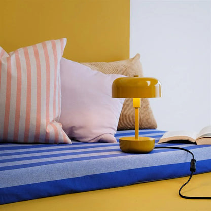 Haipot lamp in yellow, lit on a blue and yellow striped daybed, with cushions and a book.