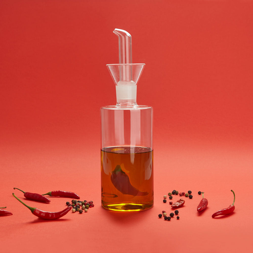 Oliera Chilli: la bottiglia di vetro con il peperoncino dentro, su sfondo rosso con peperoncini.