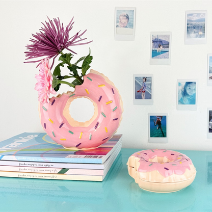 Doughnut vase in pink with flowers, resting on a stack of magazines, next to a matching trinket box.