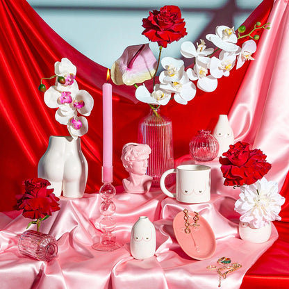 Lifestyle scene with body-shaped vases, a face mug and a bust candle on red and pink satin fabric.