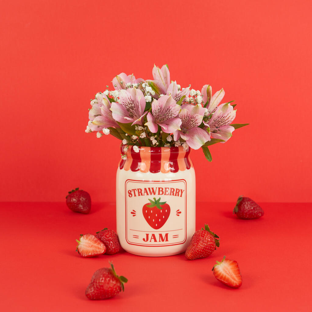 Jam vase, strawberry version with flowers, surrounded by loose strawberries on red