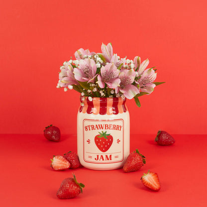 Jam vase, strawberry version with flowers, surrounded by loose strawberries on red