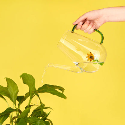 A hand pours water from the sunflower watering can onto a plant, yellow background