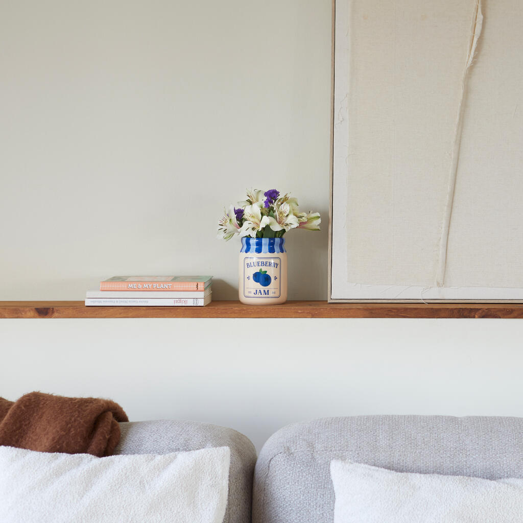 Jam vase, blueberry version on a wooden shelf, under a painting and above a grey sofa