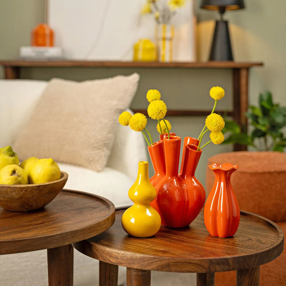 Aglio vase in orange shaped like a garlic bulb, on a side table with a yellow vase and yellow flowers.