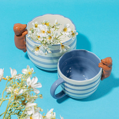 Vaso Bassotto Curioso, due tazze a righe azzurre: una con margherite, una vuota su sfondo blu.