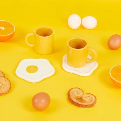 Two yellow mugs with fried-egg-shaped saucers on a yellow background with oranges and toast