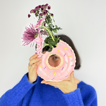 Doughnut vase in pink with flowers, held up to cover the face of someone in a blue jumper.