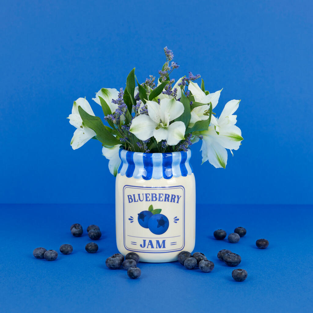 Jam vase, blueberry version with white flowers, among scattered blueberries on dark blue