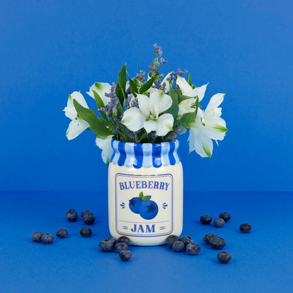 Jam vase, blueberry version with white flowers, among scattered blueberries on dark blue