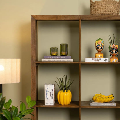 Bananas vase in yellow shaped like a bunch, on a bookshelf among head-shaped pots and a decorative bunch.